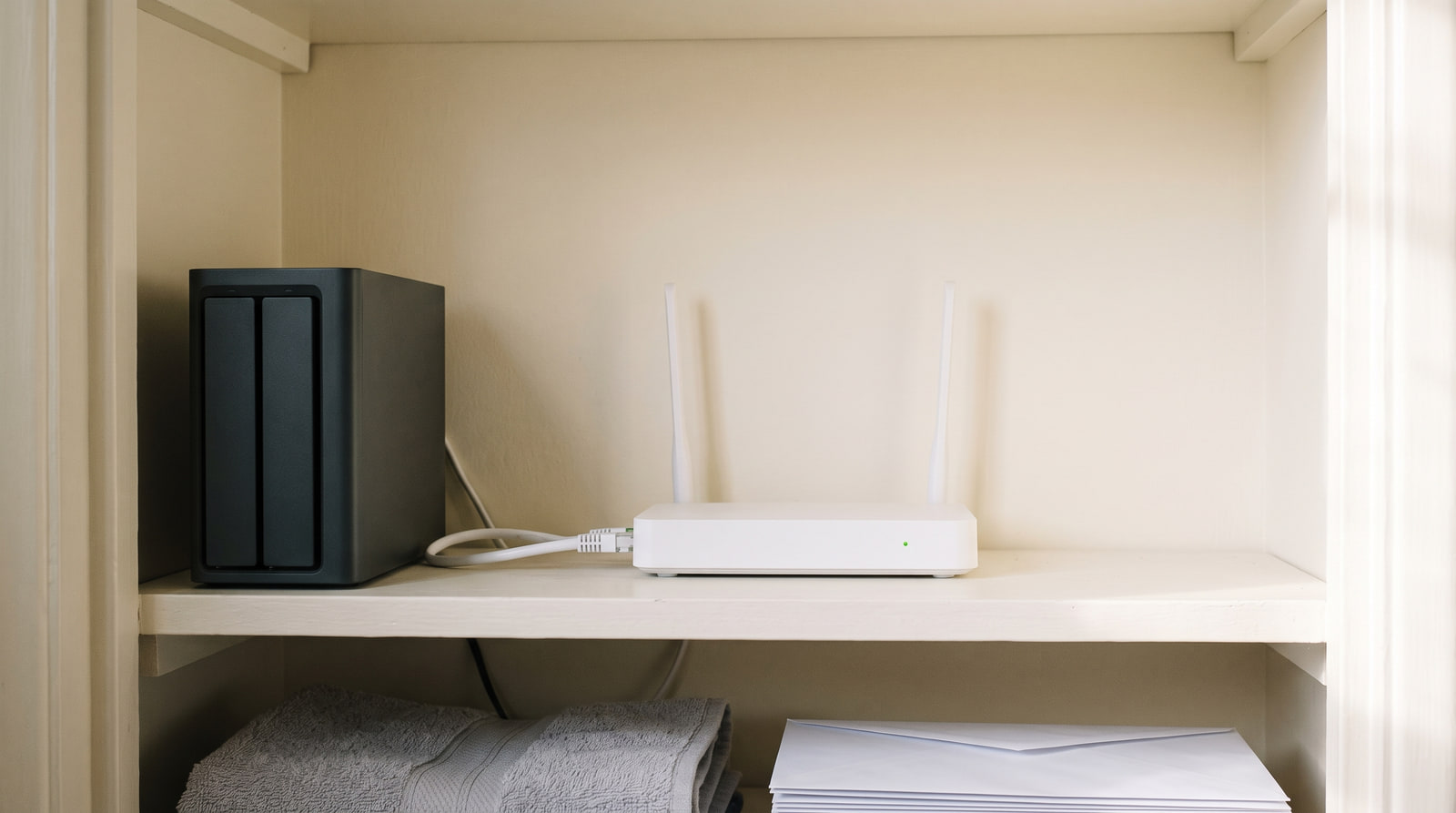 A small unbranded two-bay drive enclosure standing beside a plain white home router on a painted cupboard shelf, joined by a short ethernet cable