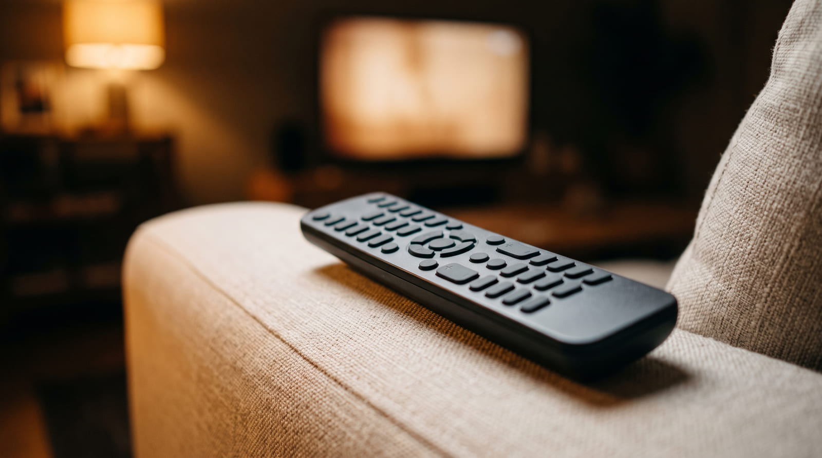 A plain remote control resting on a sofa arm with a lit television blurred behind it