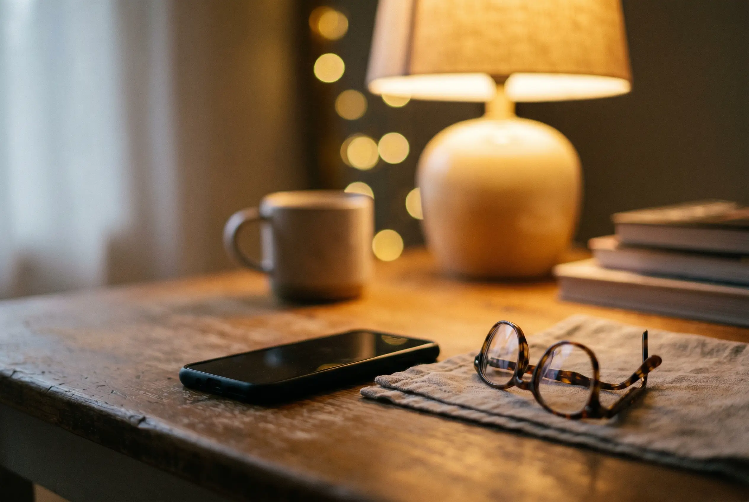 A smartphone and reading glasses on a wooden table beside a warm lamp at dusk